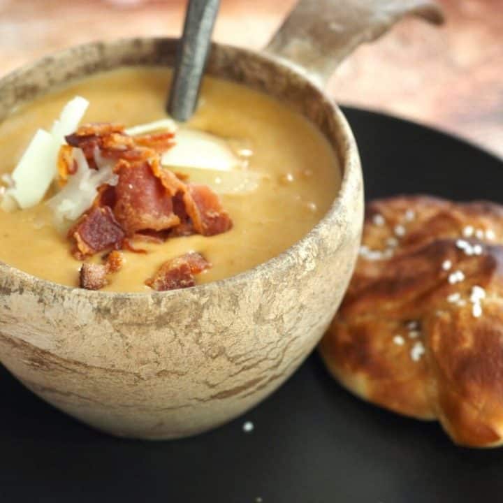 Stone bowl filled with Beer Cheese Soup, sitting on a black plate with a soft pretzel beside it.