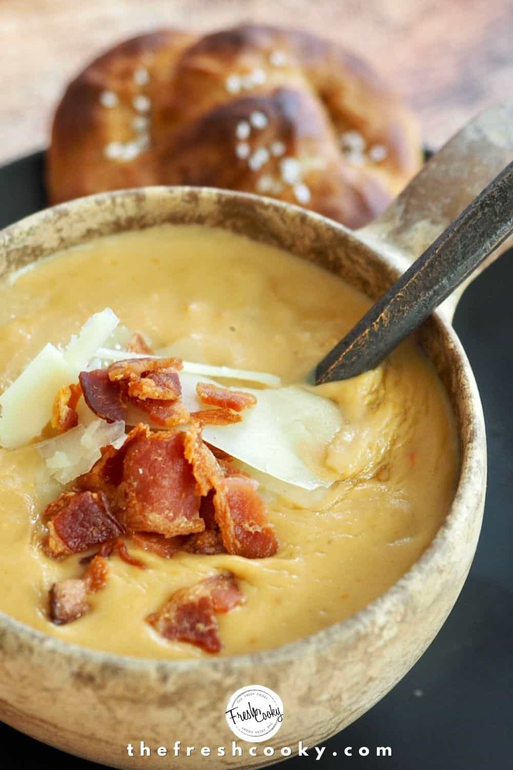 Image of a stone bowl filled with creamy, beer cheese soup, topped with shredded white cheddar and crumbled bacon, spoon in bowl with soft pretzel in background.