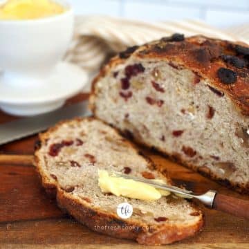 Square image of loaf of cranberry walnut bread sliced with butter being spread on the slice.