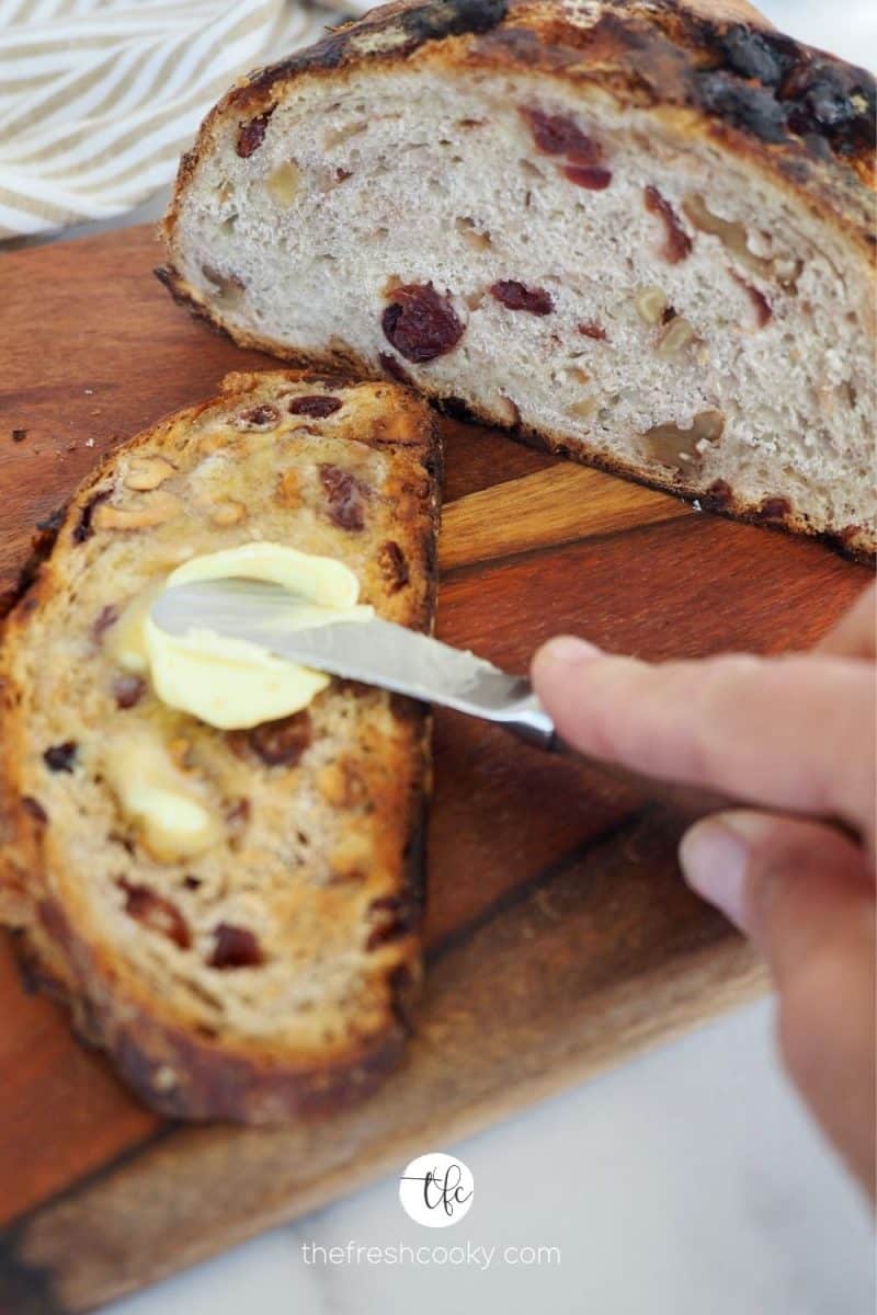 Hand spreading butter on a toasted sliced of rustic overnight cranberry walnut bread.