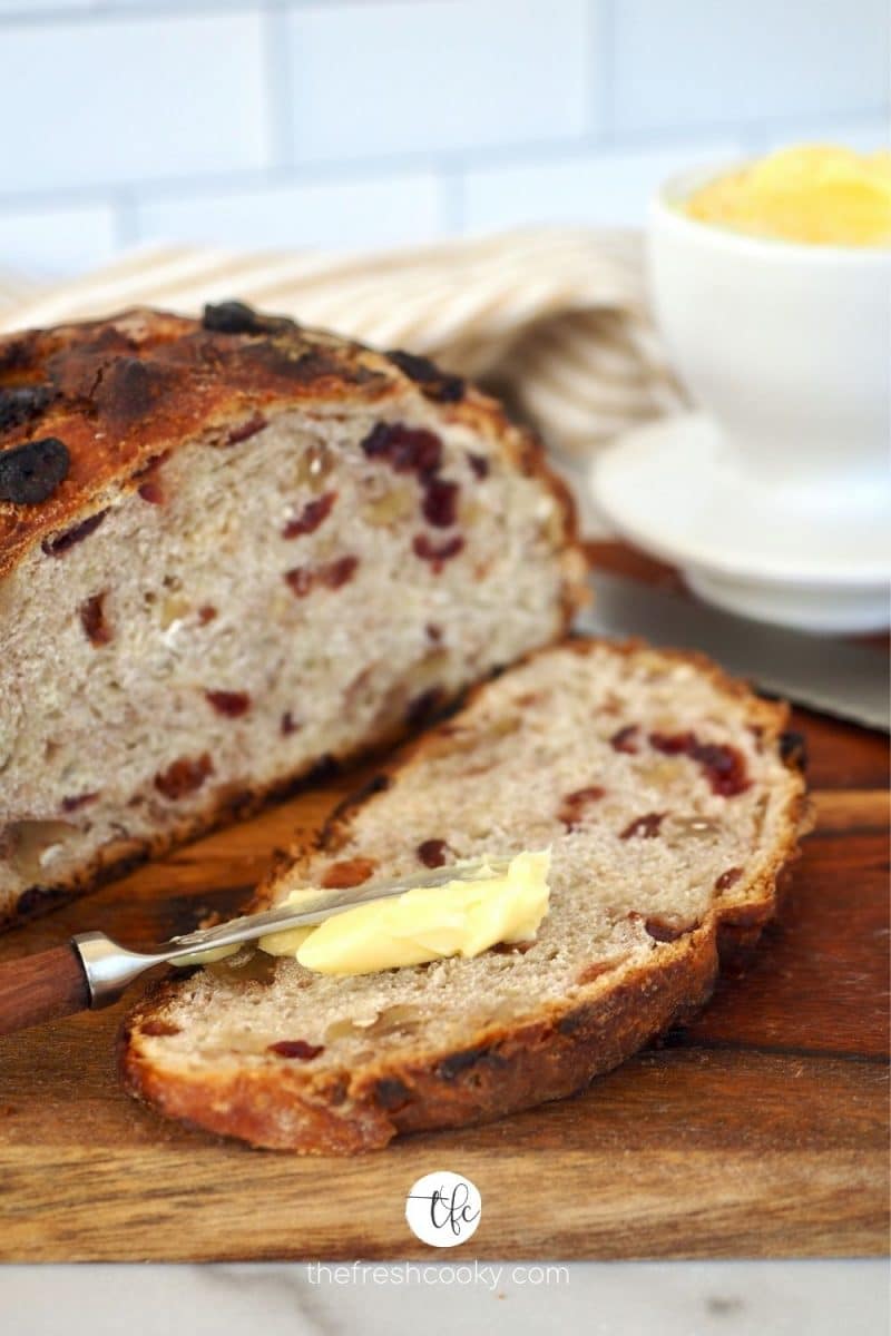 Cranberry Walnut Bread a no knead rustic overnight bread on cutting board with slice and butter knife spreading butter on slice.