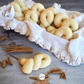 Horizontal image of Koulourakia Greek Butter Cookies in basket with ruffled napkin and one on table with smashed cinnamon sticks
