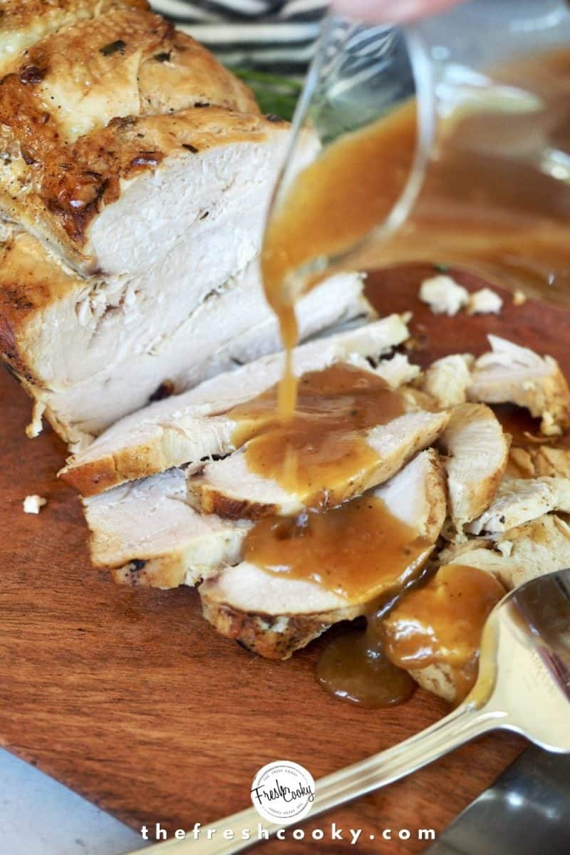 A small glass pitcher pouring turkey gravy on top of sliced turkey breast that is sitting on a cutting board, with large fork in foreground.