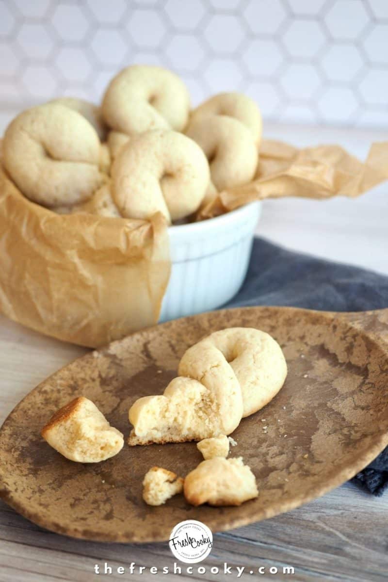 Broken Koulourakia cookie on wooden plate with small bowl lined with parchment behind stuffed with more baked cookies.