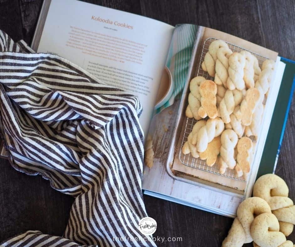 Cookbook open to recipe for Koloodia cookies with a striped tea towel laying on top and real Koulourakia cookies to the right of the cookbook