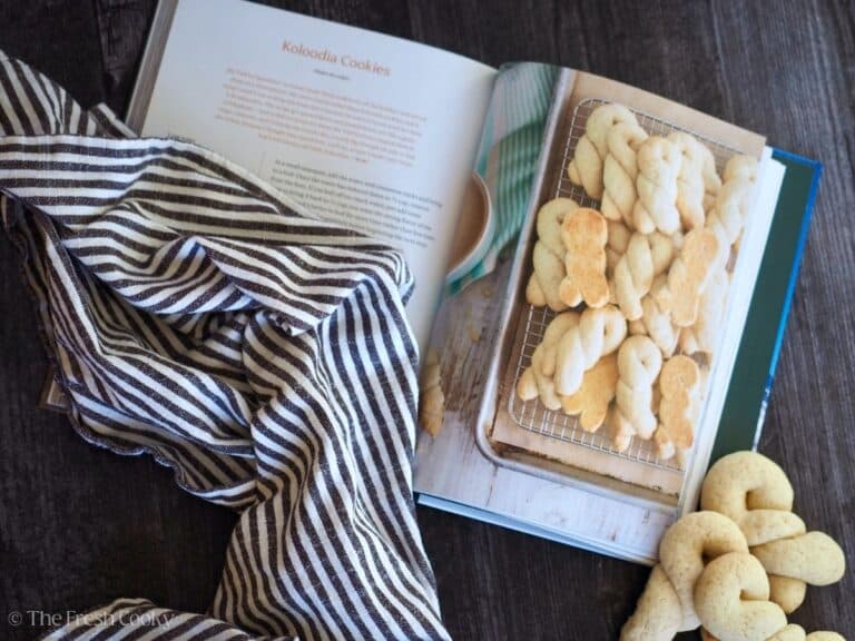 Cookbook open to recipe for Koloodia cookies with a striped tea towel laying on top and real Koulourakia cookies to the right of the cookbook.