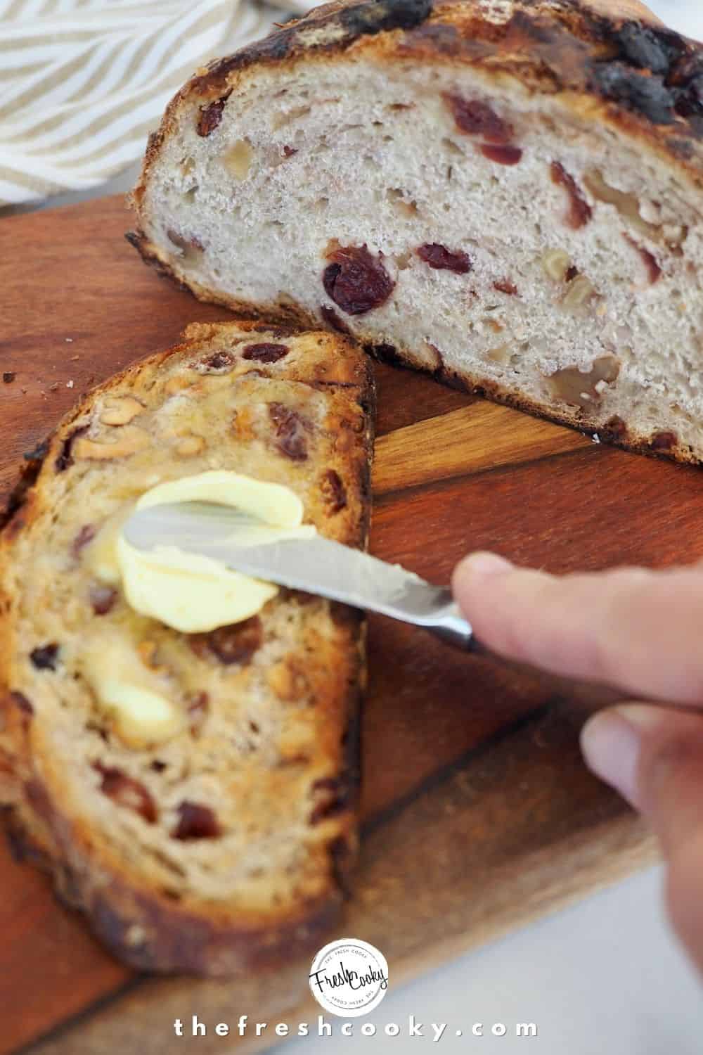 Slice of cranberry nut bread toasted, with hand buttering the slice and loaf in background.