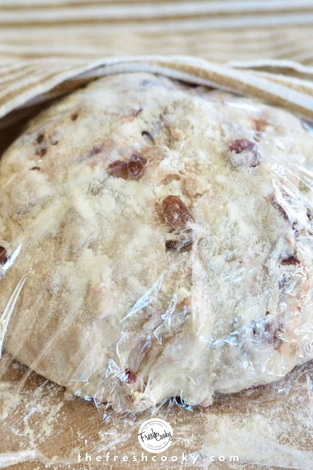 Cranberry walnut dough shaped into loaf, resting on floured parchment topped with plastic wrap, resting.