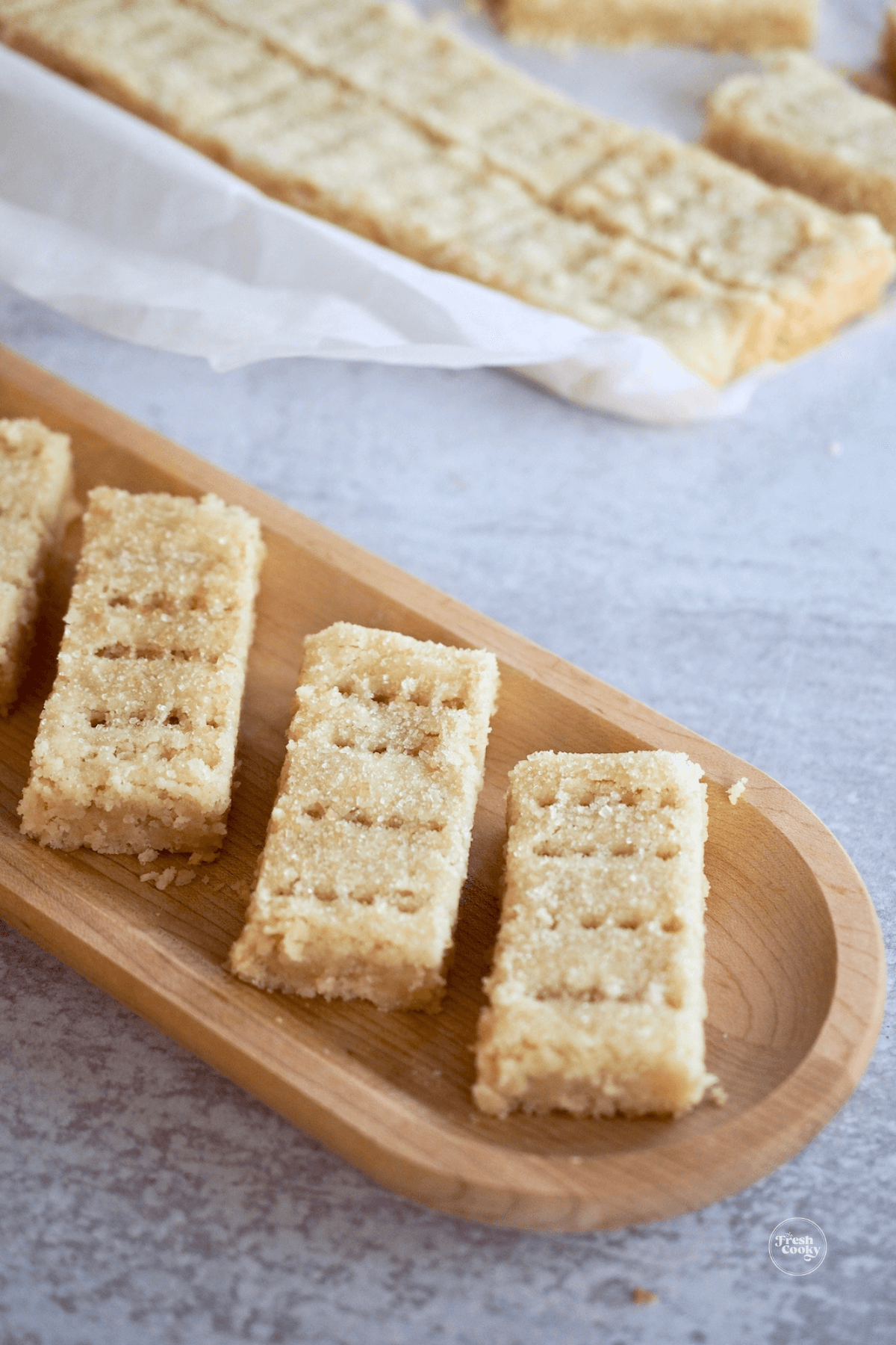 Shortbread fingers on wooden tray with more in baking pan in background.