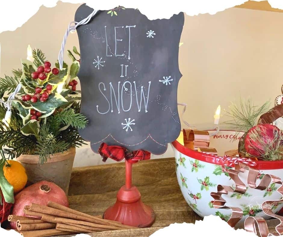 Image of Christmas Kitchen decor vignette, small potted evergreen with orange, pomegranate and cinnamon sticks, next to a small chalkboard "Let it Snow" sign, next to a Merry Christmas Batter Bowl filled with copper Christmas cookie cutters all sitting in a wooden bread board.