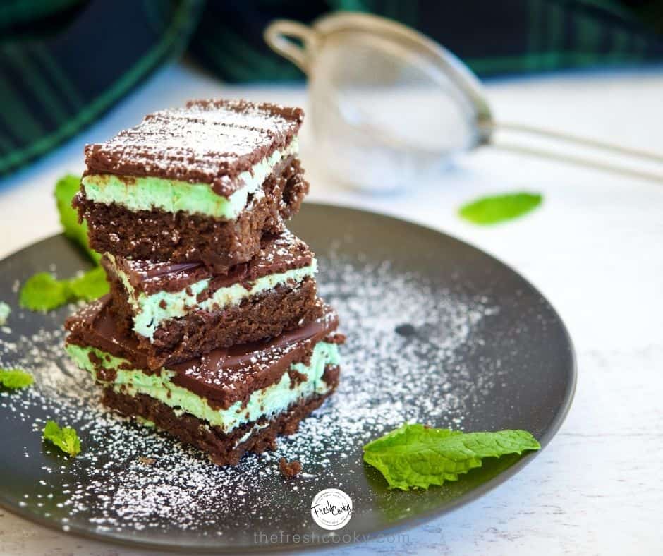 Mint Fudge Brownies on a black plate, with 3 stacked brownies, fresh mint and powdered sugar sprinkled on top