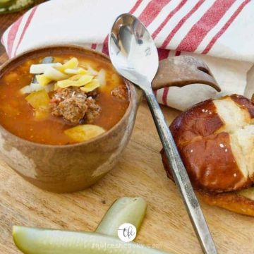 Healthy Cheeseburger Soup in a brown bowl with a rustic spoon leaning against the handle, on a wooden tray with pretzel roll and pickle spears