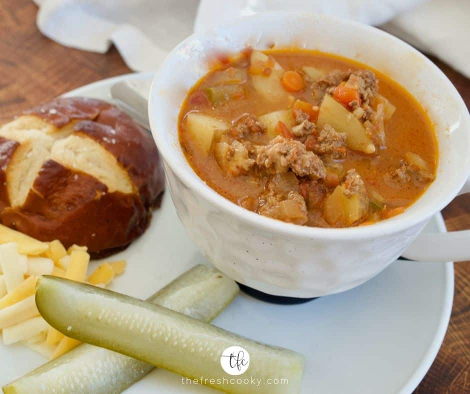 large white mug filled with healthy cheeseburger soup on a white place with pretzel roll, shredded cheese and two pickle spears.