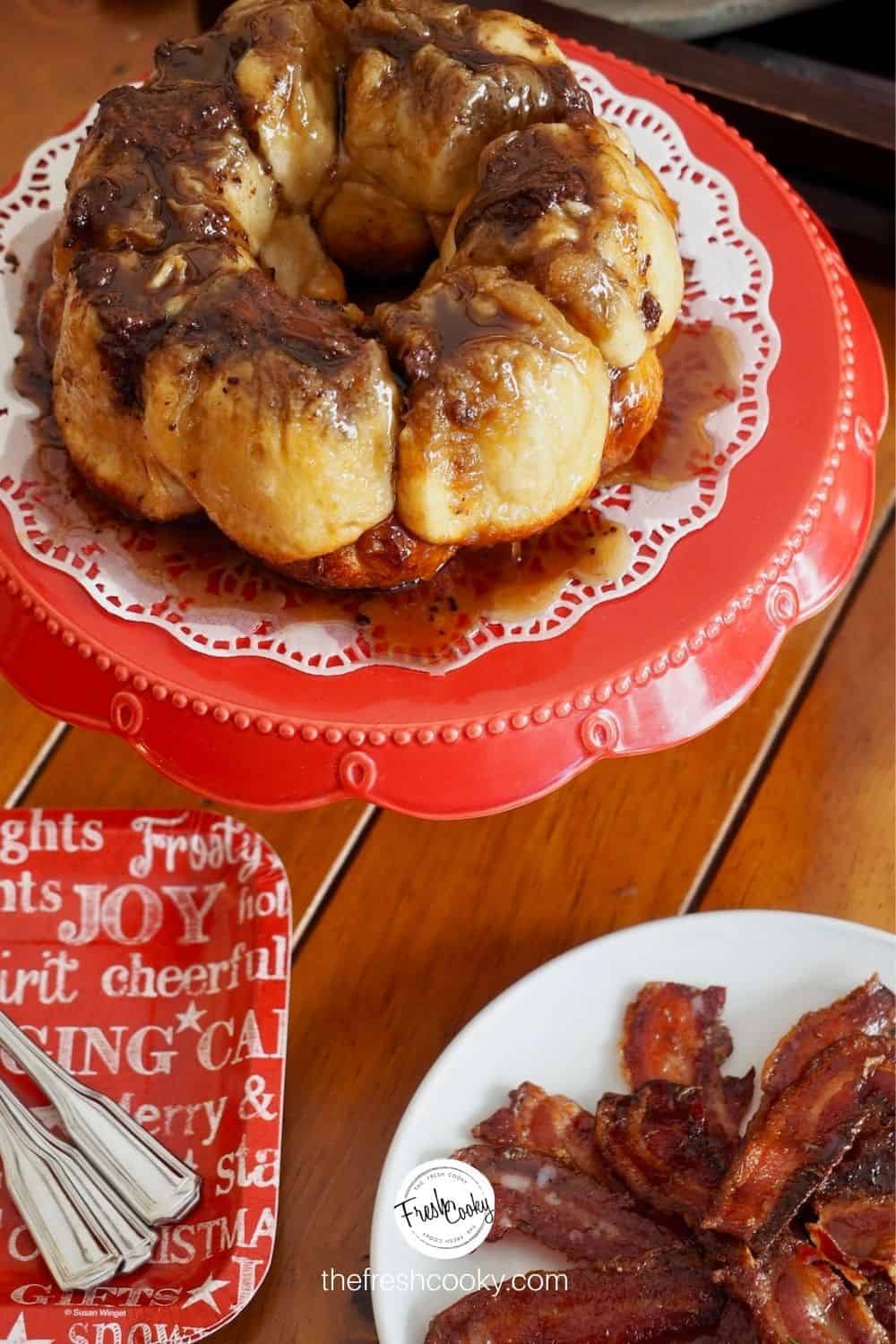 image of small chocolate monkey bread recipe on a red pedestal with a doily, sitting on a table with candied bacon and paper plates with forks nearby.