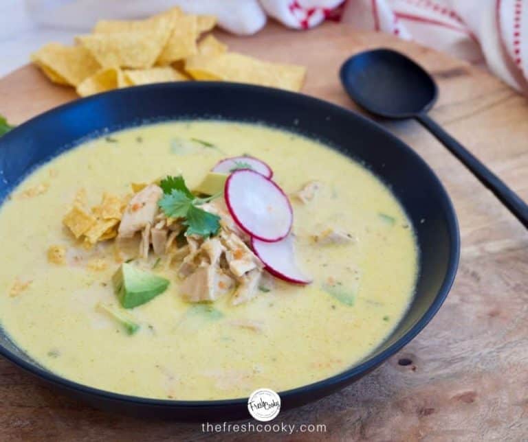 Facebook Image of black bowl of creamy chicken poblano soup garnished with avocado, radish slices, tortilla chips with a black spoon on the side and tortilla chips in the background.