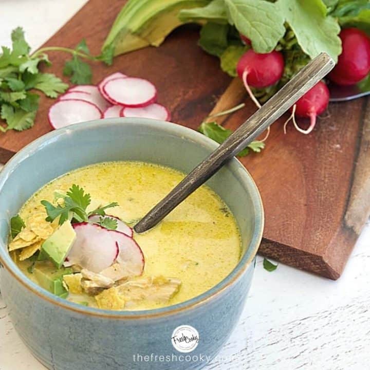 Facebook image for Creamy Chicken Poblano Soup in a rustic clay blue bowl with rustic soup spoon. with radishes on cutting board, fresh cilantro