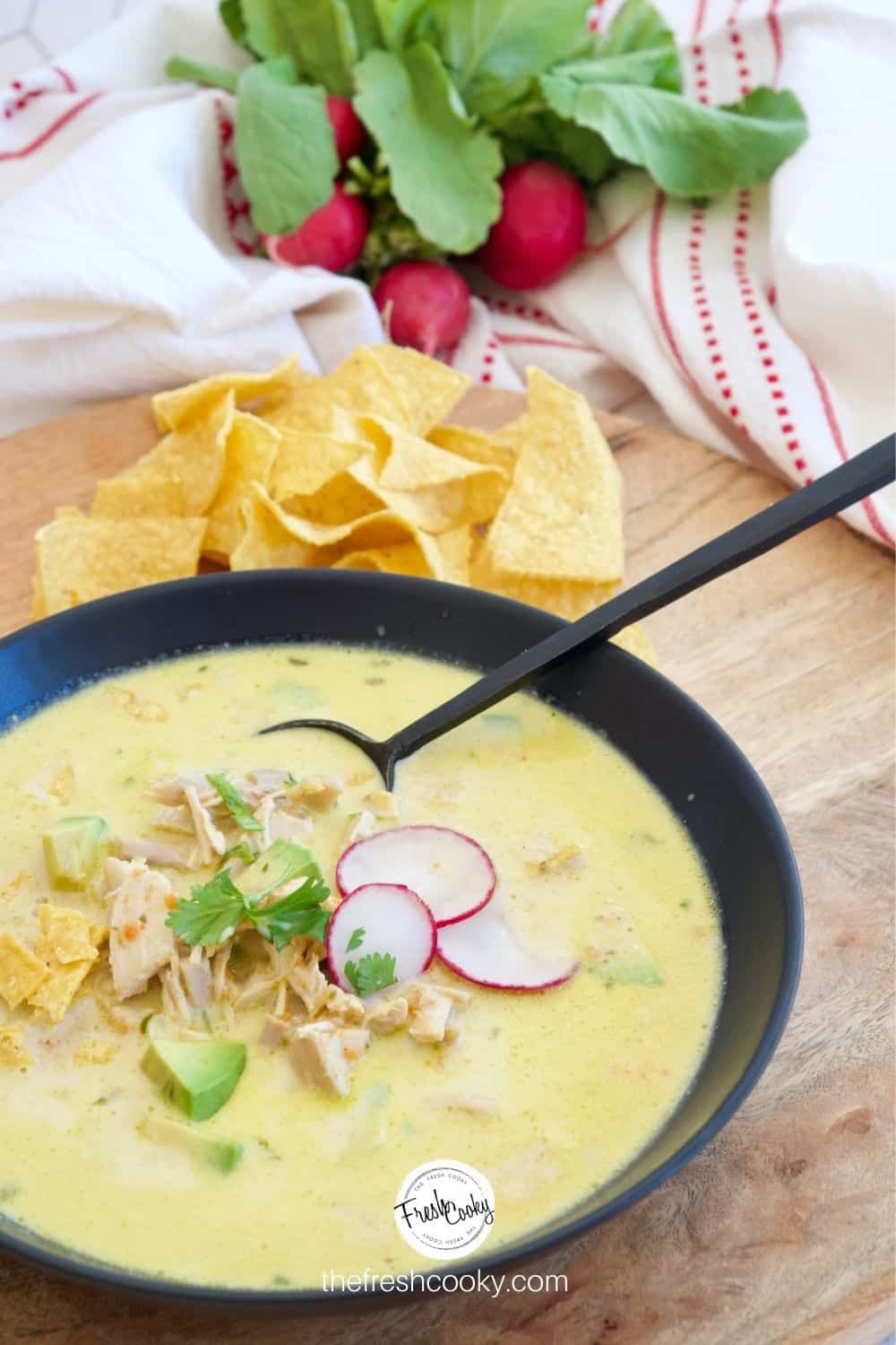 Creamy Chicken Poblano Soup in a black bowl with a black spoon on a wooden tray with tortilla chips and radishes in the background.