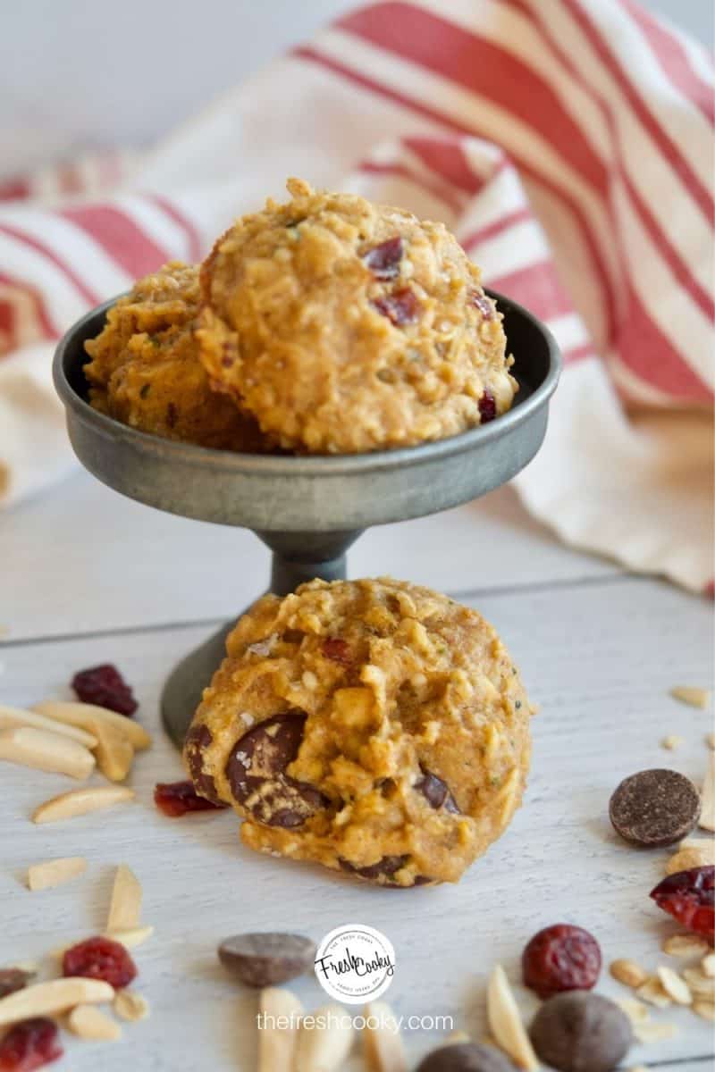 Gluten Free Breakfast Cookies image with two cookies sitting on a galvanized pedestal and one cookie at the base with dried craisins, almonds and chocolate chips and a striped napkin in the background.