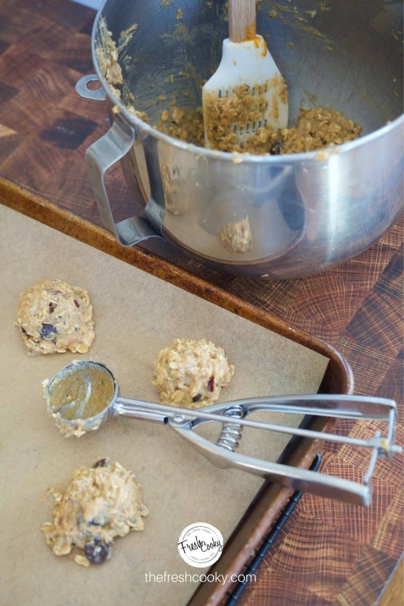 Image of cookie sheet, lined with parchment paper and medium cookie scoop on tray with scooped Gluten Free Breakfast Cookies on tray, mixing bowl in background.