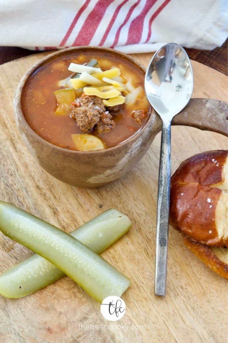 Wooden bowl with handle filled with healthy cheeseburger soup topped with farm cut cheesee, pretzels, dill pickle spears on a wooden tray