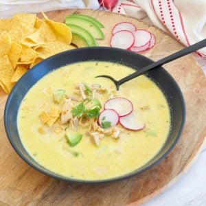 Creamy Chicken Poblano Soup recipe in black bowl garnished with radishes, avocado and tortilla chips.
