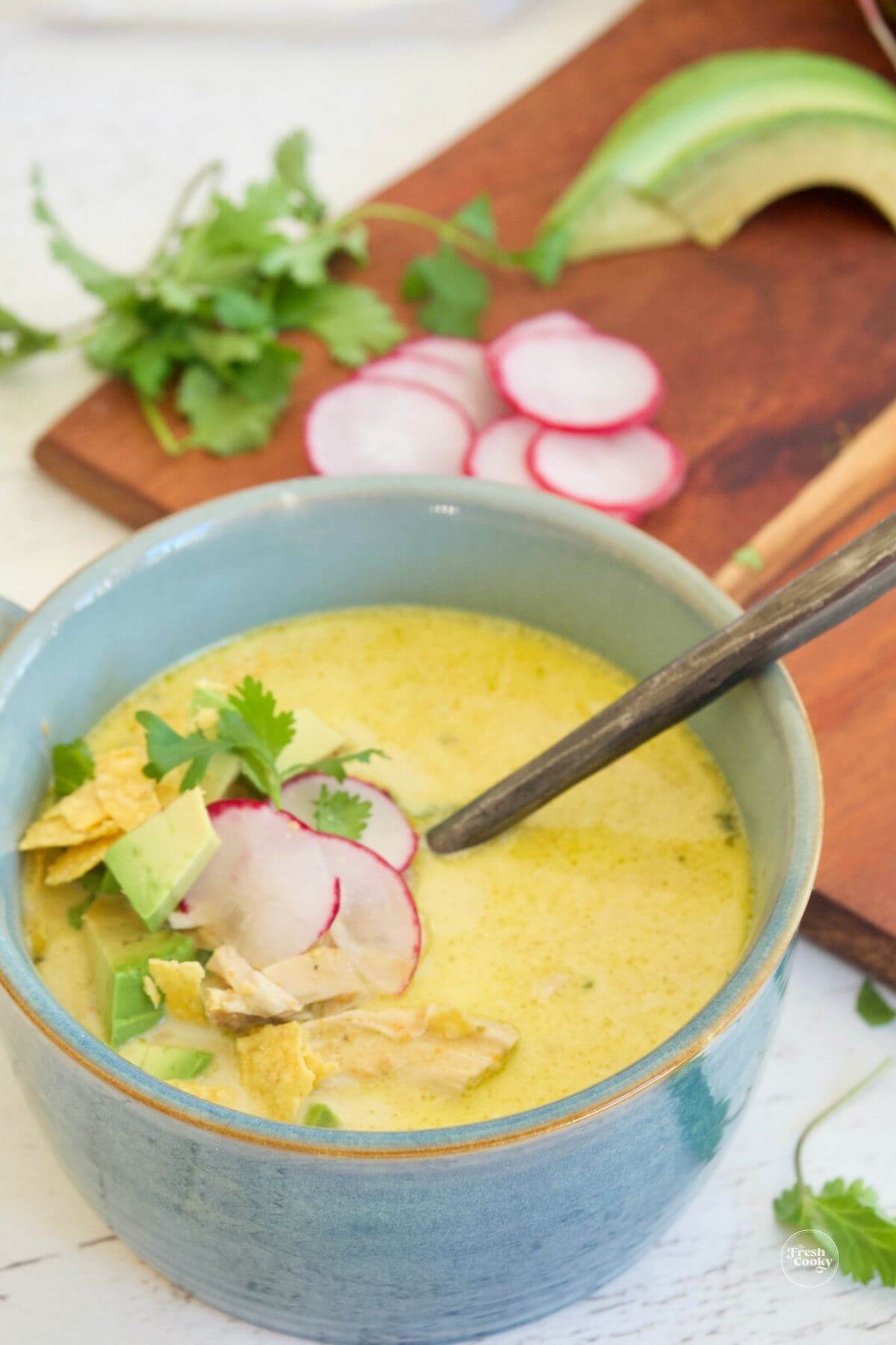 Blue bowl filled with yellow tinted chicken poblano soup garnished with radishes and avocados.