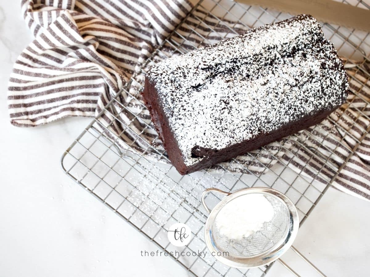 Easy chocolate loaf cake dusted with powdered sugar, sitting on a wire rack with a striped towel beneath and powdered sugar in a small sieve sitting beside.