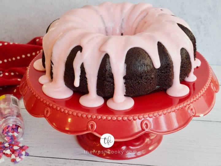 Horizontal image of easy chocolate loaf cake in bundt form, drizzled with puddles of pink glaze, with sprinkles on the side.