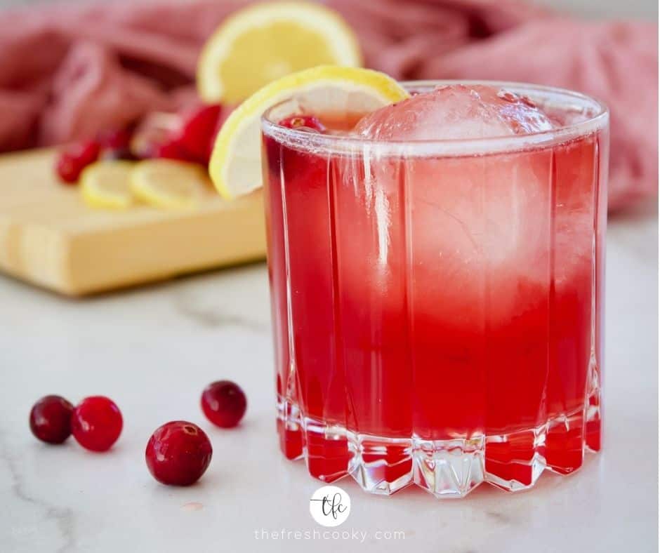 Cranberry Bourbon Sour facebook image with sphere ice cube, garnished with a lemon wedge, pink napkin in background frozen cranberries on marble by the cocktail