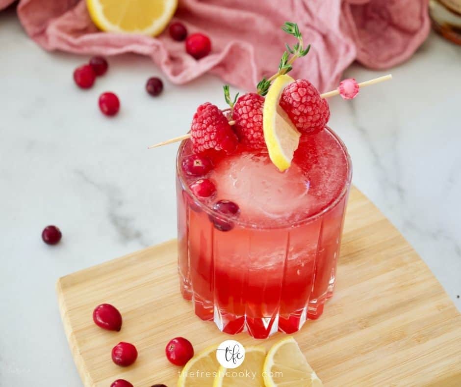 Facebook image of Cranberry Bourbon Sour on mini cutting board with cranberries and lemon slices on top, garnished with raspberries in a cocktail pick and a sprig of thyme, a pink napkin in the background