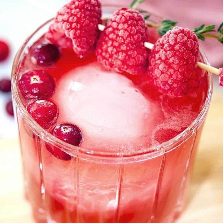crystal glass filled with cranberry bourbon sour, garnished with large sphere ice cube, cranberries and pick with three frozen raspberries