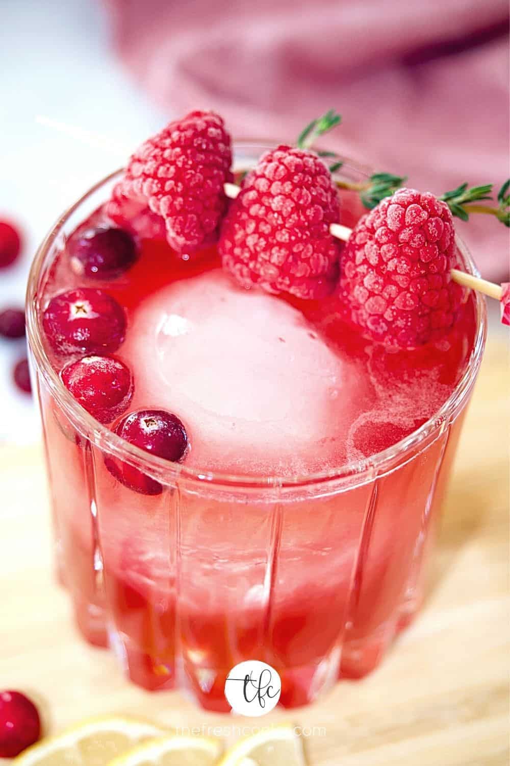 Crystal glass filled with cranberry bourbon sour, garnished with large sphere ice cube, cranberries and pick with three frozen raspberries.