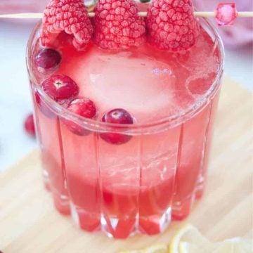 Cranberry Bourbon Sour in crystal glass on wood cutting board with cranberries and frozen raspberries on a cocktail pick garnish.