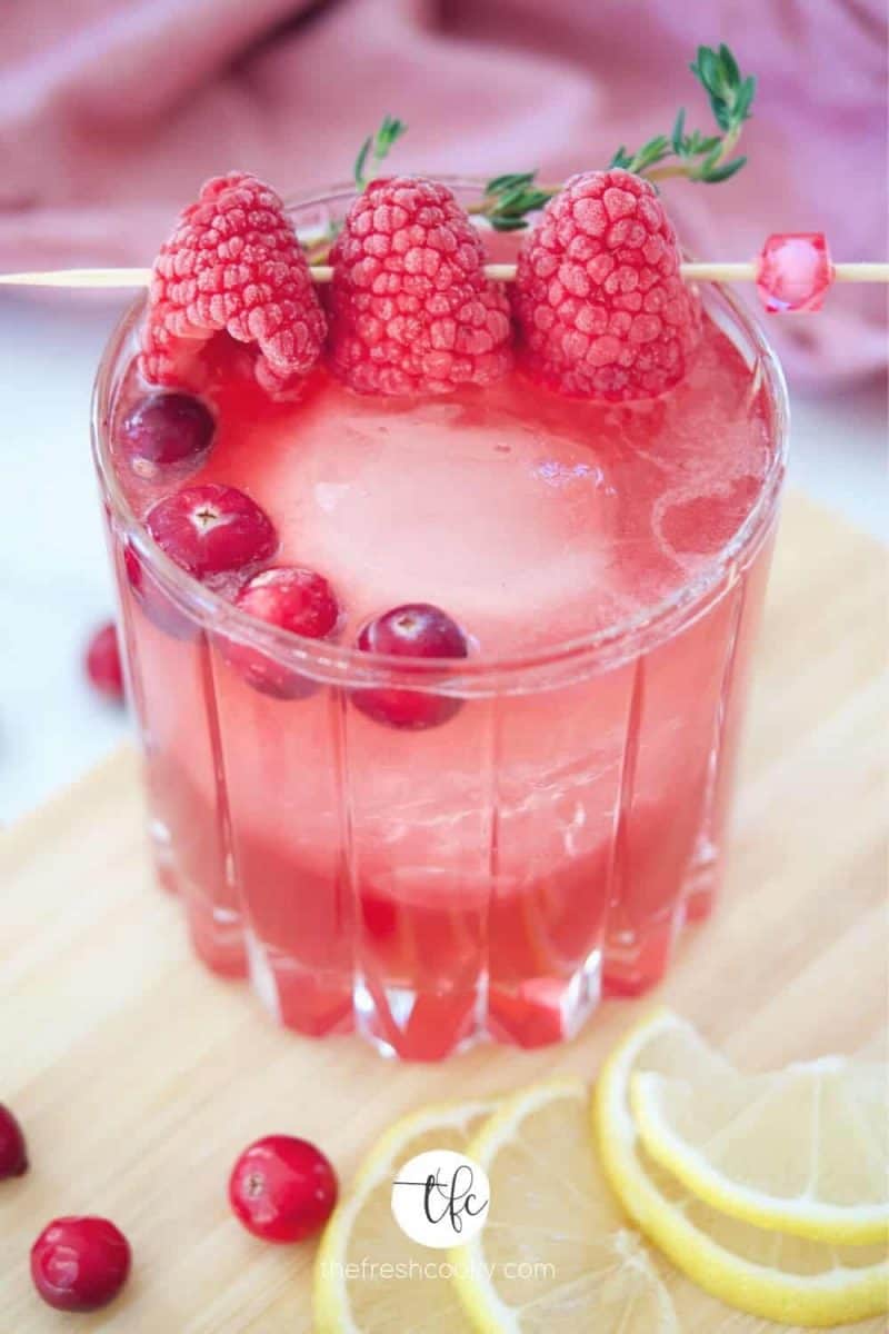 Cranberry Bourbon Sour in crystal glass on wood cutting board with cranberries and frozen raspberries on a cocktail pick garnish.