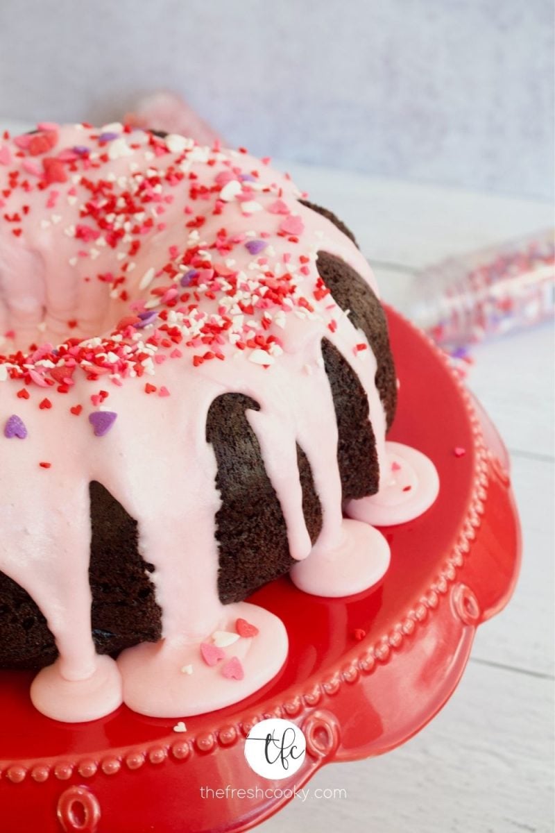 chocolate bundt cake covered with pink vanilla buttercream and heart sprinkles on a red pedestal plate