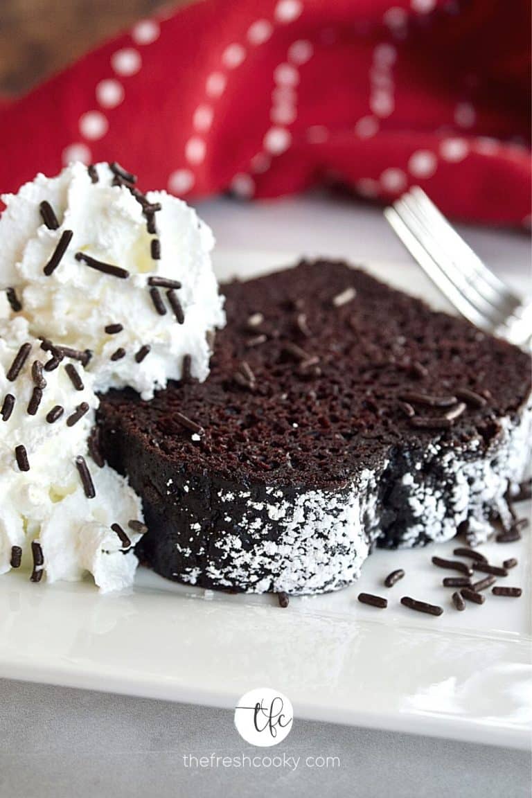 image of slice of easy chocolate loaf cake on white plate with fork, dusted with powdered sugar and whipped cream with chocolate jimmies, red napkin in background