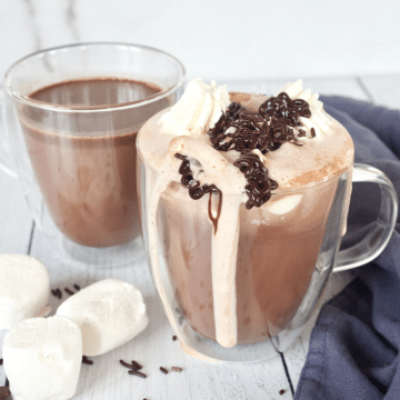 Homemade Hot Chocolate with chocolate chips in pretty double walled glass mugs filled with whipped cream and drizzled with chocolate.