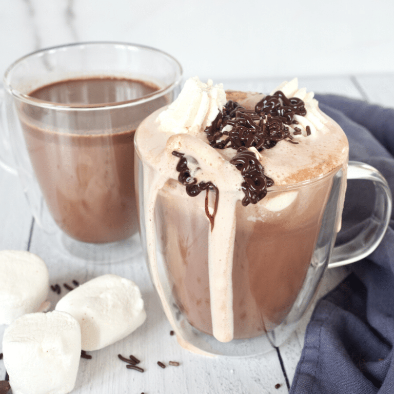 Homemade Hot Chocolate with chocolate chips in pretty double walled glass mugs filled with whipped cream and drizzled with chocolate.