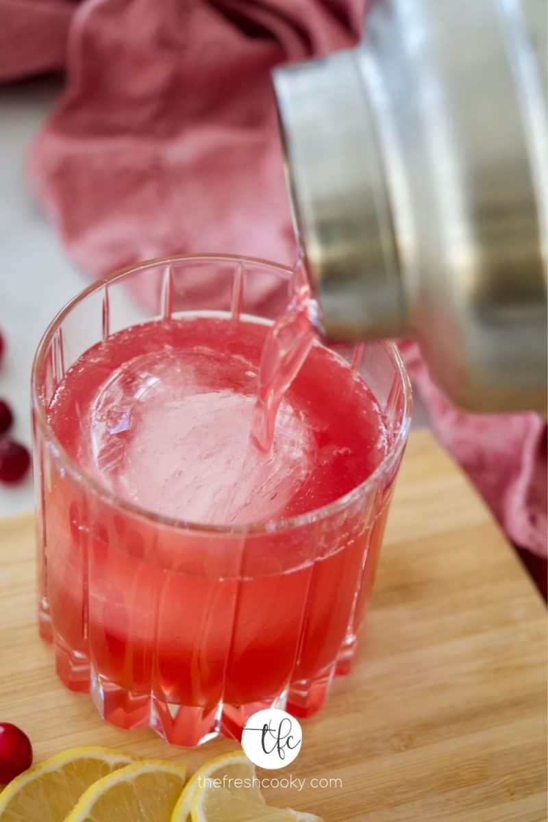 Pouring cranberry bourbon sour cocktail from shaker into crystal glass with sphere ice cube