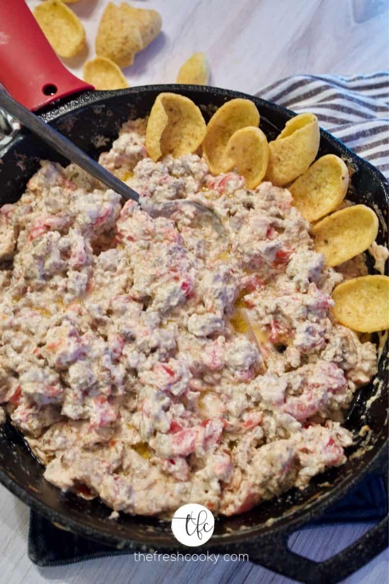 cast iron skillet filled with sausage cream cheese dip, lined with frito scoops on one side with a silicone handle on pan, striped towel in background