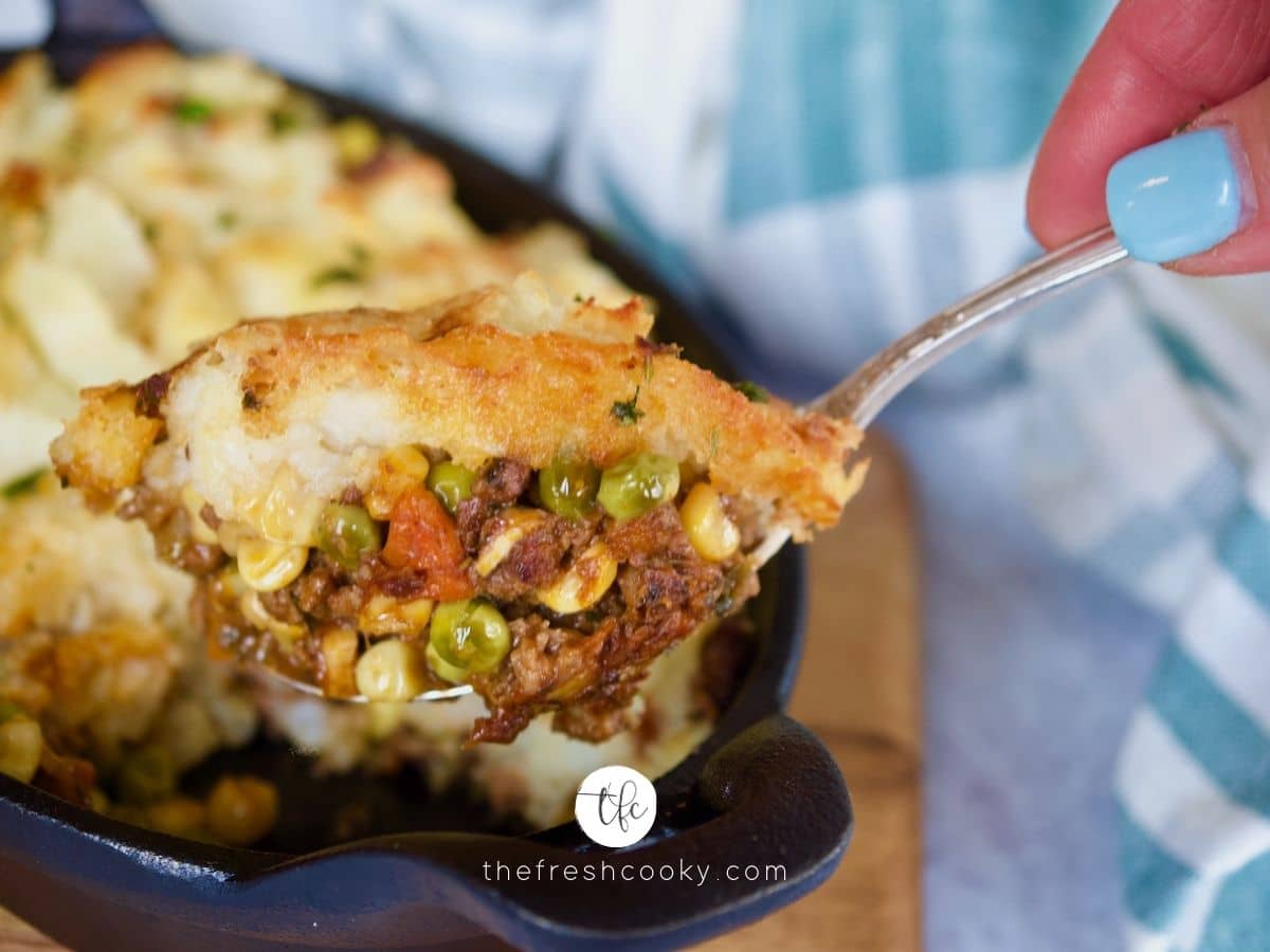 cast iron pan filled with skillet shepherd's pie with hands bringing up spoonful.