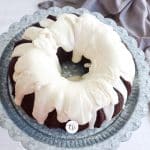 landscape image of chocolate bundt cake drizzled with vanilla buttercream glaze on pedestal with gray tea towel