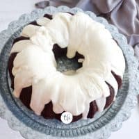landscape image of chocolate bundt cake drizzled with vanilla buttercream glaze on pedestal with gray tea towel