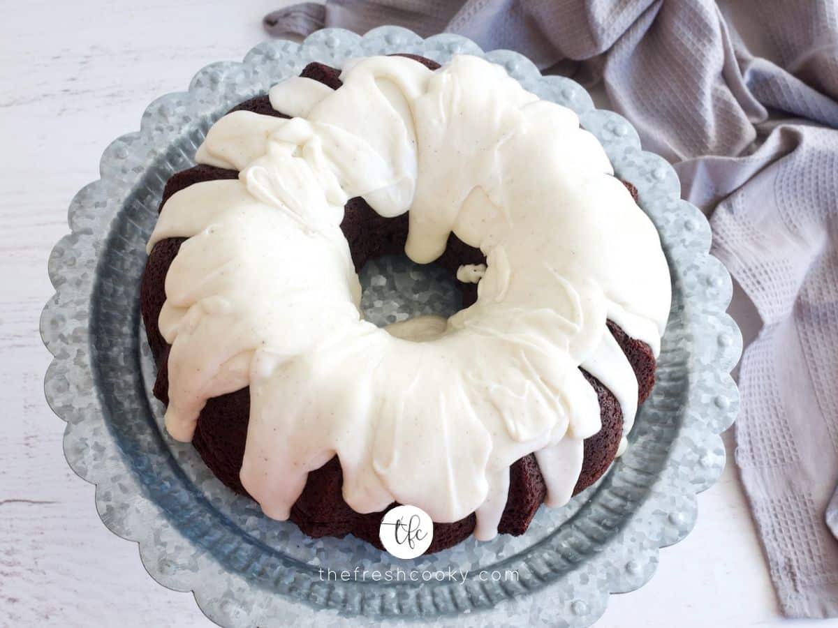 landscape image of chocolate bundt cake drizzled with vanilla buttercream glaze on pedestal with gray tea towel