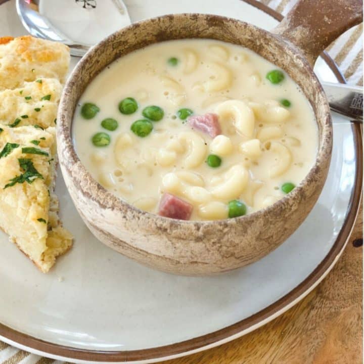 Stoneware bowl full of Ham, Mac & Cheese Soup with wedge of biscuit on wooden charger.