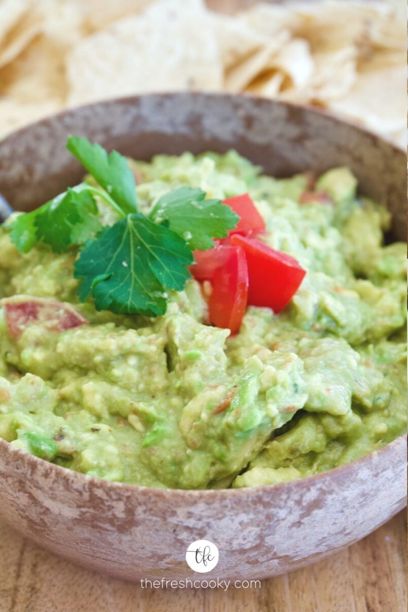 Large wooden bowl filled with bright green fresh guacamole with cilantro and tomatoes and tortilla chips in background.