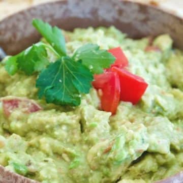 Large wooden bowl filled with bright green fresh guacamole with cilantro and tomatoes and tortilla chips in background.