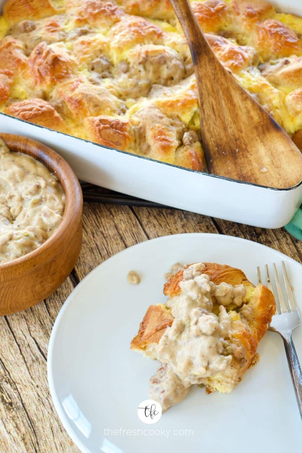 a slice of biscuits and gravy egg bake on plate with pan behind, smothered in sausage gravy, with bowl of gravy on the side.