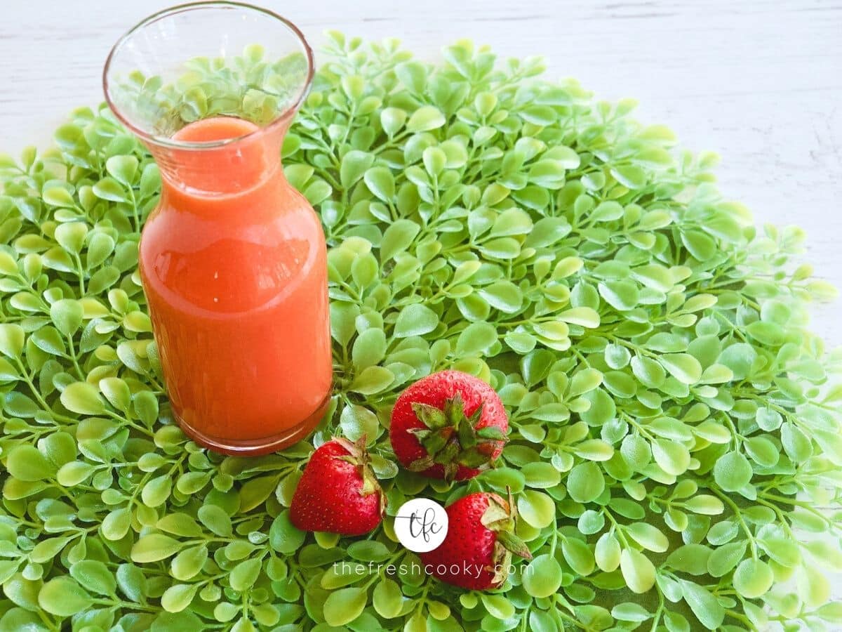 Fresh Strawberry Syrup in jar with three strawberries nearby, sitting on bed of greens.