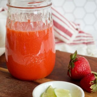 A mason jar of fresh strawberry syrup on a cutting board with strawberries and lime by the syrup.